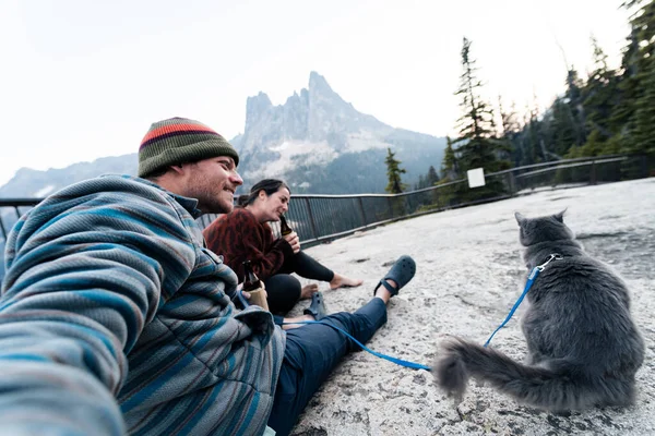 Young 30s caucasian woman sits with her husband and cat enjoying a drink while having her gray russian blue cat on leash in wilderness forest while at popular road trip rest area in washington state.