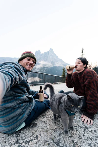 Young 30s caucasian woman sits with her husband and cat enjoying a drink while having her gray russian blue cat on leash in wilderness forest while at popular road trip rest area in washington state.