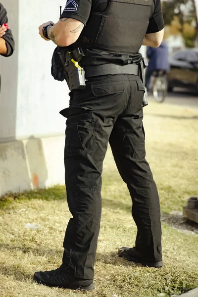 White male police officer cop standing on grass in a park during the day talking with person. Cop holding something in hand. Wearing bullet proof jacket and taser on left hip. Shoulder and below.
