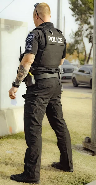 White male police officer cop standing on grass in a park during the day talking with person. Cop holding something in hand. Wearing bullet proof jacket and taser on left hip.