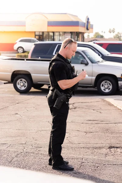 White male caucasian police officer cop trooper standing on street with black uniform and gun on hip during the day writing down information on potential suspect in a bad part of the city.