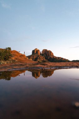 Young beautiful woman standing on rock point looking out at Cathedral Rock in Sedona Arizona USA Southwest with incredible sunset colors and beautiful reflection from small pool of water on red stone.