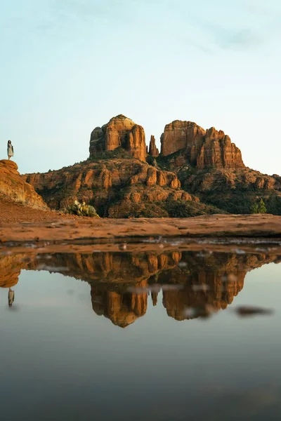 Young beautiful woman standing on rock point looking out at Cathedral Rock in Sedona Arizona USA Southwest with incredible sunset colors and beautiful reflection from small pool of water on red stone.