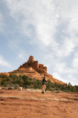Woman hiker standing on Bell Rock trail in red rock formations within coconino national forest in Sedona Arizona USA against white cloud background.