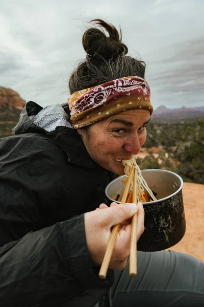 30 'lu yaşlarındaki güzel beyaz bir kadın Sedona Arizona' da yemek çubuklarıyla kamp yerinden ramen yerken kameraya gülümsüyor.