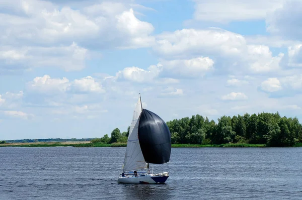 Lovely landscape with one small sailboat with black and white sails, which smoothly slips on the water surface of the lake on a summer day against the green shore and the blue sky with beautiful clouds.