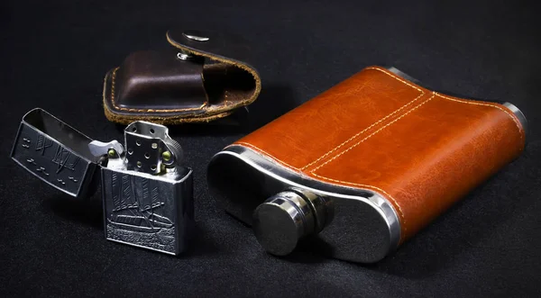 Alcoholic flask in leather trim with an old-fashioned petrol lighter and a leather case on a black background. Close-up