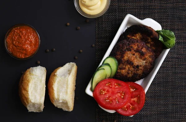 Cutlets with vegetables in a white plate with containers of sauces under dark lighting. Shallow depth of field