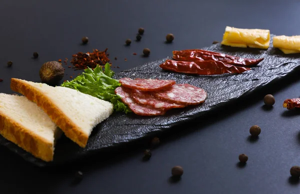 stock image Sliced smoked salami sausage with bread, vegetables and other ingredients on a black stand. Shallow depth of field