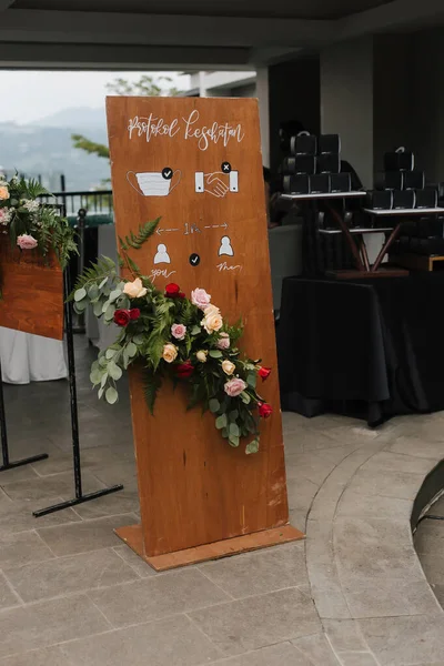 A front board sign translate in Indonesia say: Health protocol, with flower decoration standing at the wedding entrance.