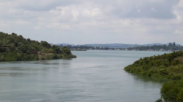 stock image A large river with green trees on the left and right ended into a lake with a mountain background in the horizon with bright cloudy sky.