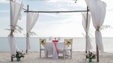 Romantik yemek masası seti ve beyaz temalı sandalye hazırlığı, beyaz kum, açık bulutlu gökyüzü ve sakin, huzurlu, güzel sahil manzaralı deniz manzarası. Evlilik teklifi için iyi bir yer.