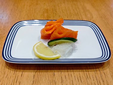 Mavi çizgili beyaz dikdörtgen bir tabakta taze somon balığı, limon dilimi ve jülyen salatalıkla süslenmiş, suşi restoranındaki ahşap bir masaya karşı.