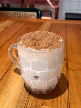 Buzlu çikolatalı içecek, üzerinde kakao tozu ve tahta bir kafeterya masasında cam bardak.