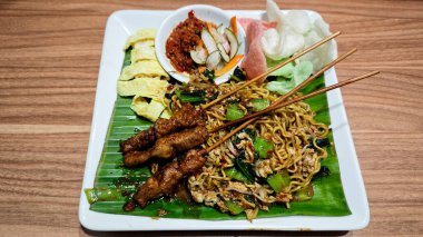 Izgara tavuk şişleri, omlet dilimleri ve muz yapraklı tabakta sambal ve krakerlerle servis edilen karışık sebzeler ahşap bir masada sunuluyor.