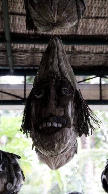 Bali Kültür Köyü 'nün geleneksel bambu çatısından sarkan el oyması korkunç yüzlü Rustik Hindistan cevizi kabuğu heykeli.