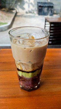 Layered Avocado Mocha Drink with Ube and Sweet Toppings in a Glass on a Wooden Table in an Outdoor Setting clipart