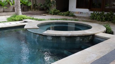 Lüks açık hava jakuzisi ve berrak, güneşli bir günde tropik bir tatil köyünde yemyeşil ve modern villa elementleriyle çevrili sakin mavi fayanslarla dolu yüzme havuzu..