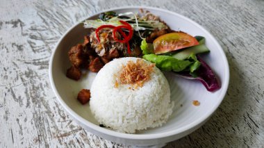 Kızarmış domuz eti, pilav ve taze Sambal Matah. Köhne ahşap bir masada salata garnitürle servis ediliyor.