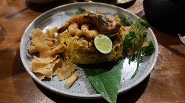 Limonlu bir tabakta servis edilen yumurta ağı garnitürlü deniz ürünleri Char Kway Teow ve bir restoranda ahşap masa üzerinde gevrek krakerler