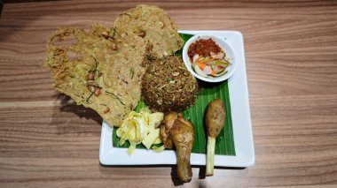 Traditional Indonesian Fried Rice with Sambal and Crispy Peanut Crackers on Banana Leaf Plating with Grilled Chicken Drumstick and Sate Lilit Garnished on White Square Plate clipart