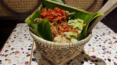 Geleneksel Nasi Bakul kırmızı biberli sambal, kızarmış arpacık soğanı ve örülmüş bambu sepetinde servis edilen ve renkli bir terrazzo masasında muz yapraklarıyla süslenmiş Endonezya yemeği.