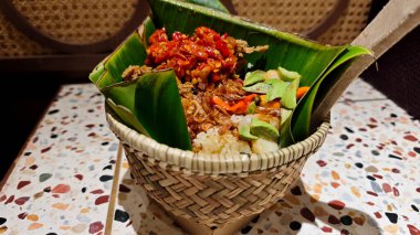 Traditional Nasi Bakul with red chili sambal, fried shallots, and petai served in a woven bamboo basket lined with banana leaves on a colorful terrazzo table in an Indonesian dining setting clipart