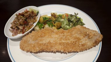 Ekmek arası balık filetosu sotelenmiş biberli soğan kavrulmuş patates taze yeşil salata ve maydanozlu garnitür beyaz tabakta canlı ve iştah açıcı bir yemek sunumu yaratır..