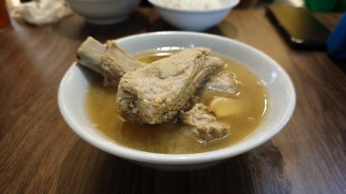 Beyaz bir seramik kasede servis edilen sarımsak ve hafif et suyu ile buharlı bir kase domuz pirzolası çorbası ahşap bir masada, bulanık bir arka planla günlük yemek düzeninde.