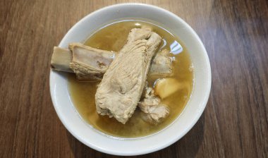 Beyaz bir seramik kasede servis edilen sarımsak ve hafif et suyu ile buharlı bir kase domuz pirzolası çorbası ahşap bir masada, bulanık bir arka planla günlük yemek düzeninde.