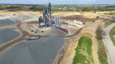 Asfalt bitkisi, uzun depolama siloları ve büyük kum ve çakıl yığınları etrafında çalışan ağır makineleri olan bir çakıl ocağının havadan görünüşü. Sanayi sahasında altyapı için toplam çıkartma, asfalt üretimi ve malzeme arzı vurgulanıyor