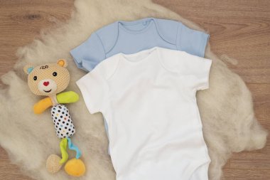 Ahşap arka planda beyaz ve mavi bebek tulumu modeli ve renkli ayı oyuncağı. Boş bebek kıyafetleri maket gibi. Düz yatmış stok fotoğrafı.
