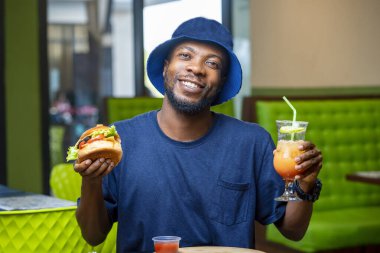 image of excited african guy holding glass of juice and sandwich- black guy enjoying snacks in a restaurant
