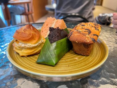 Tabakta lezzetli hamur işleri sergileniyor: bir altın-kahverengi kremalı pasta, bir poğaça, bir kremalı kek, bir dilim kremalı pasta, ve muz yapraklı çikolatalı brownie kek..