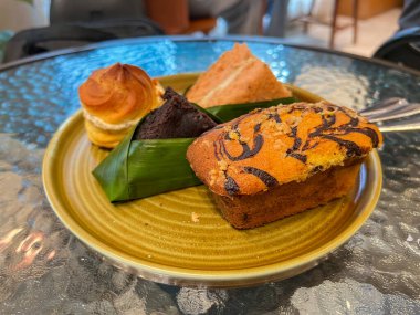 Tabakta lezzetli hamur işleri sergileniyor: bir altın-kahverengi kremalı pasta, bir poğaça, bir kremalı kek, bir dilim kremalı pasta, ve muz yapraklı çikolatalı brownie kek..