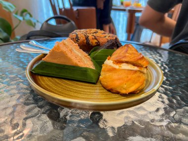 Tabakta lezzetli hamur işleri sergileniyor: bir altın-kahverengi kremalı pasta, bir poğaça, bir kremalı kek, bir dilim kremalı pasta, ve muz yapraklı çikolatalı brownie kek..