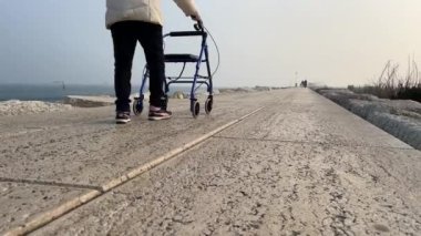 Old woman walks on the seashore with a walker