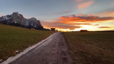 Moving along road in mud in front of mountain and on green meadow at sunset
