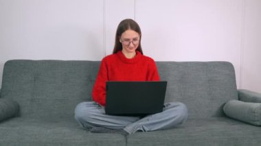 Kanepede oturan gözlüklü kadın diz üstü bilgisayar kullanarak bacak bacak üstüne attı. Evde dizüstü bilgisayar kullanmak, ekrana bakmak, okumak ya da e-posta yazmak, koltukta oturmak, ciddi bir öğrencinin ödevlerini yapması.,