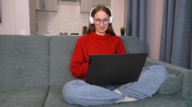 Kulaklıklı genç bir kadın dizüstü bilgisayarla konferans görüşmeleri yapıyor, online öğretmenle konuşuyor, çalışıyor, evden çalışıyor. Bilgisayar kamerası kullanarak öğrenen bayan öğrenci notlar alır. uzaklık eğitimi