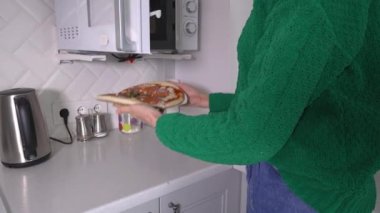 Yeşil kazaklı güzel, beyaz bir kadın mikrodalga fırına pizza koyar. Yakın plan. Modern fırında donmuş fast food pizzasını mutfak dekoruna karşı ısıtıyor..