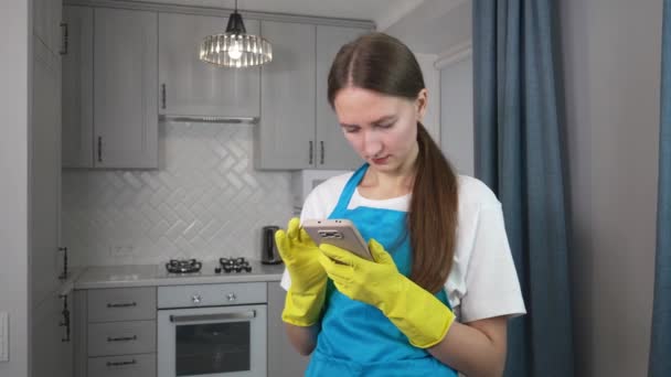 制服を着た美しい白人女性は スマートフォンのキッチンクリーニングを使用します 現代のキッチンで清掃サービスチーム メイドが携帯電話でチャット Auペアはアプリを通じてクライアントを検索します スマートホーム — ストック動画
