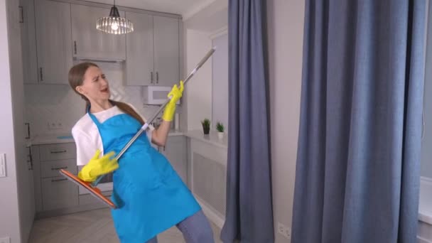 身着围裙和白色T恤的白种人女喜剧演员在度假时清扫她家厨房时 与拖把共舞 唱着滑稽的歌曲和动作 清洁公司 — 图库视频影像