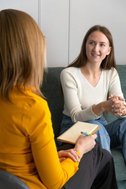 Smiling young woman listens to a psychologist at the reception, sitting on the couch in the office.Psychotherapist advises the patient about the results of the examination, gives recommendations clipart