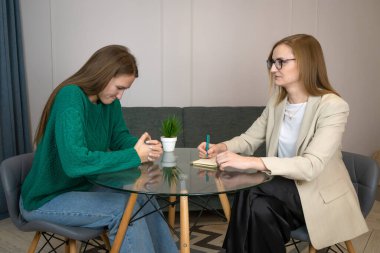 Kadın psikolog, üzgün genç bir kadınla cam bir masada resepsiyon veriyor ve defterine notlar yazıyor. Psikolojik danışmanlık.