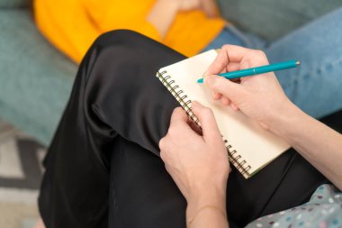 Kanepede genç bir kadınla psikoterapi seansı sırasında not alan bir psikoloğun elinde kalemle bir defterin yakın çekimi. Psikoloji, psikoterapi, ilgisizlik, yardım, terapi.