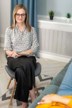 Gözlüklü, bluzlu ve siyah pantolonlu kadın psikoloğu ofisinde kanepede yatan genç bir kadınla psikolojik bir seans düzenliyor. Bir psikoterapist randevusunda. Dikey fotoğraf