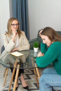 Kafası aşağıda ve dudakları birbirine yapışmış genç bir kadın bir psikolog randevusunda oturuyor. Modern bir ofiste bir psikologun danışmanlığı. Dikey fotoğraf. Seçici odak