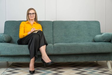 Güzel genç psikoterapist notlar alır, kameraya bakar ve koltukta otururken gülümser. Gözlüklü güzel kadın psikolog, odada pano ile poz veriyor.