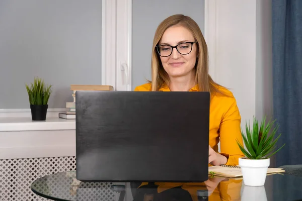 Close-up of a woman with a slight smile in glasses, an orange blouse, working at a laptop, sitting at a glass table. Concept of business, psychology, marketing, training, development.